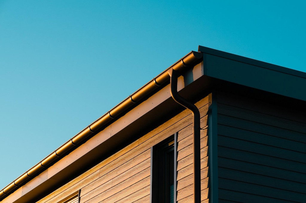 Dark seamless gutter and downspout along the roofline of a home with wood-look siding against a clear blue sky