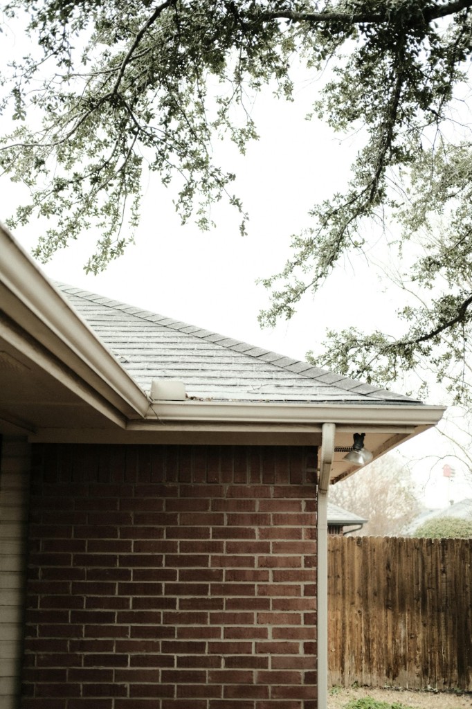 Brick home with white gutters and a downspout under oak tree branches beside a wooden fence