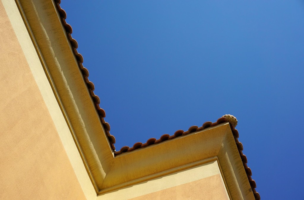 Looking up at the corner of a cream stucco home with terracotta roof tiles against a clear blue sky