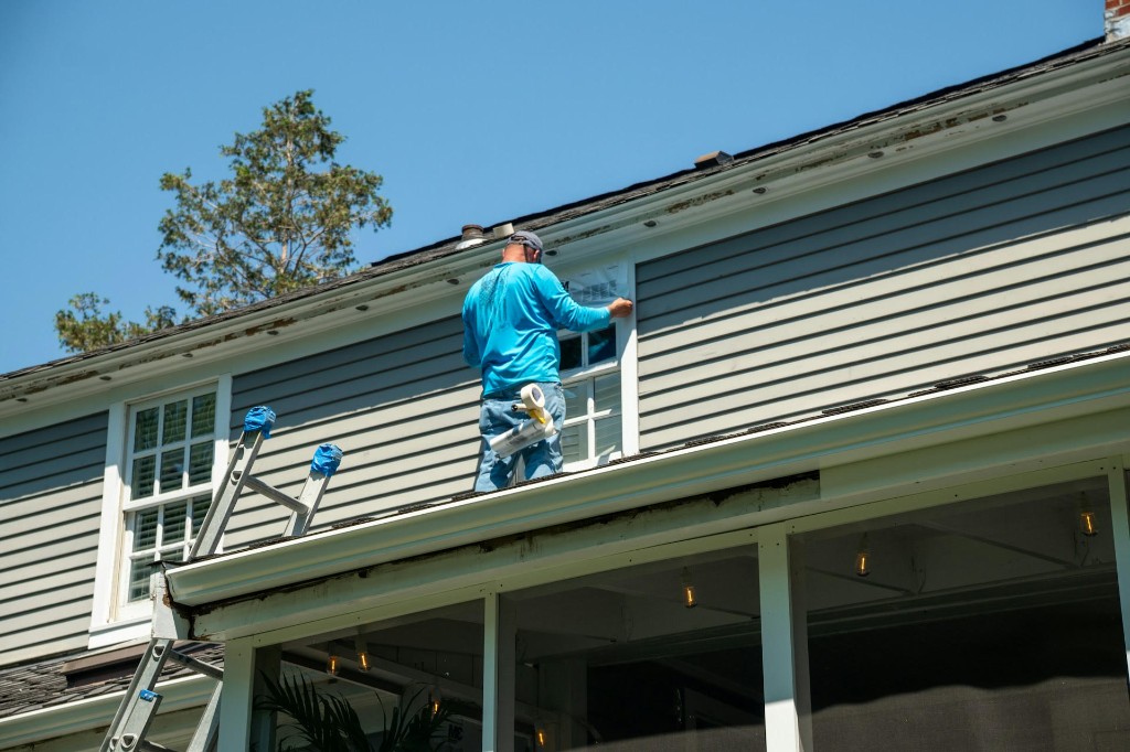 Technician on a residential roof working near white gutters and peeling fascia trim